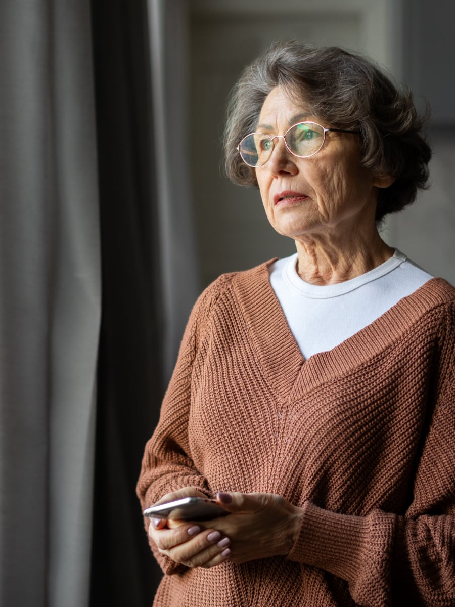older woman holding cell phone and looking out window with a contemplative look