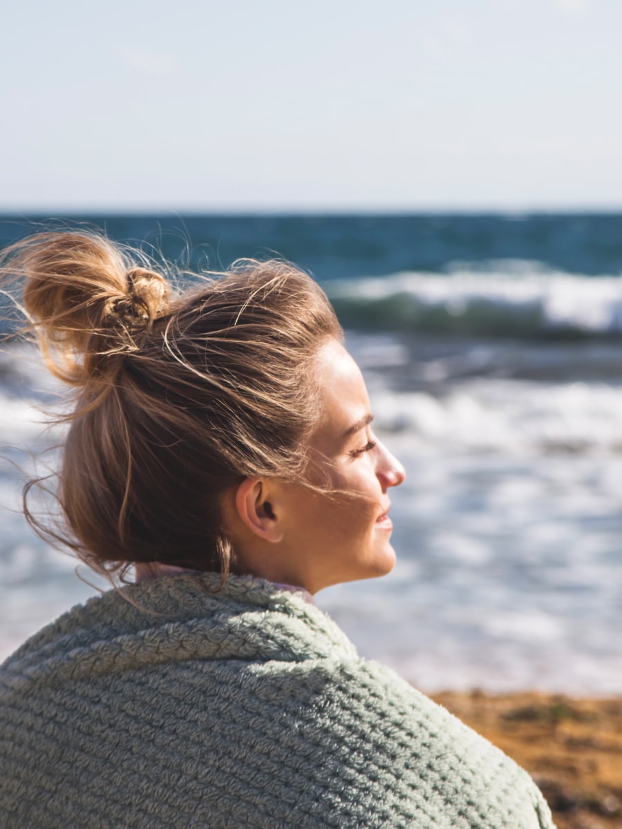woman with messy bun hair wrapped in a blanket on a beach staring off into the distance