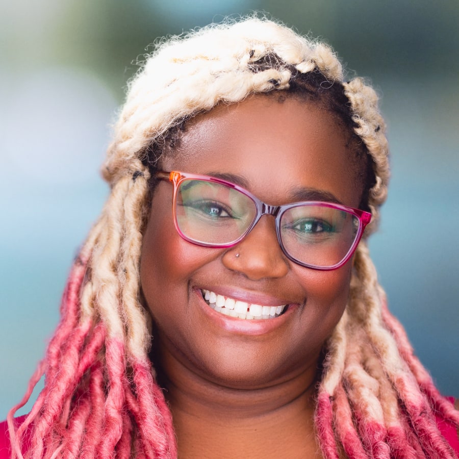 Headshot of Imaria Hardon. A young woman with blonde and pink dreadlocks and pink rimmed glasses