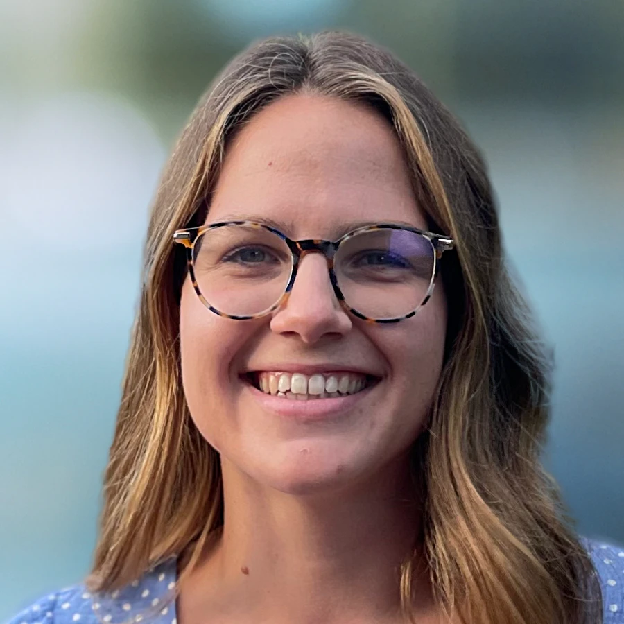 Anna Scholcz, wearing tortoiseshell glasses and a blue shirt, smiles widely at the camera. She has long blonde hair against a blurred outdoor background.